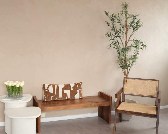 A wooden bench and a woven chair are positioned beside a small table and a vase of white tulips against a beige wall.