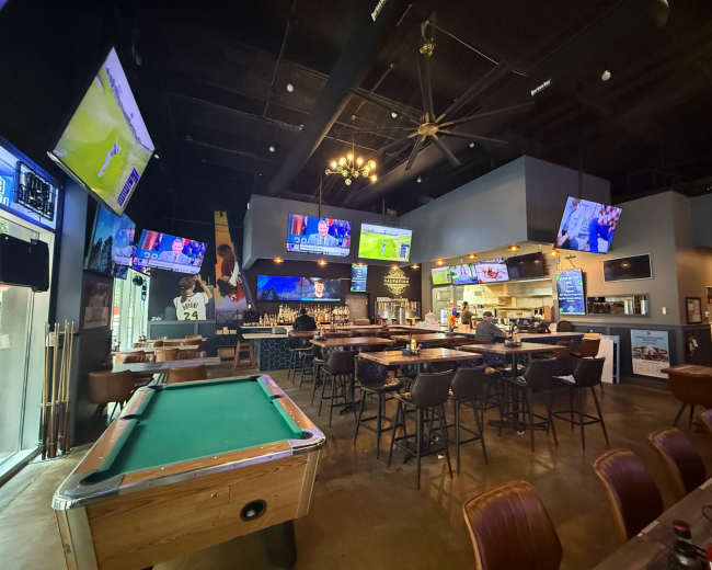 The image shows an interior of a bar or restaurant featuring several televisions, a pool table, and a mix of high-top and standard seating.