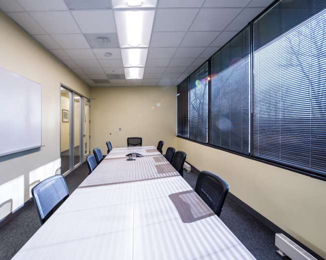 A long meeting table with several black chairs is set up in a brightly lit conference room with large windows.
