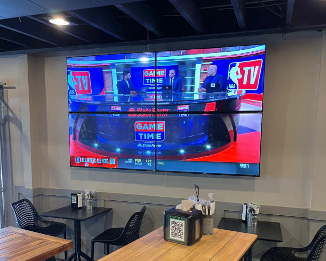 A large flat-screen television displays a sports broadcast in a dining area with wooden tables and black chairs.