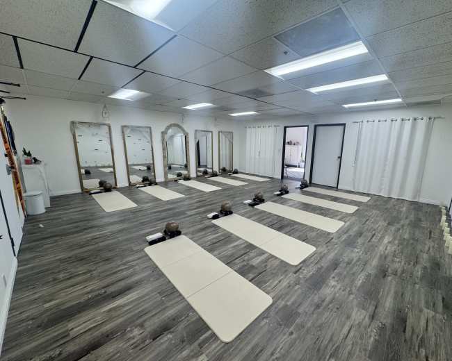 The image shows a spacious yoga studio with several exercise mats arranged neatly on the floor, surrounded by mirrors and natural lighting.