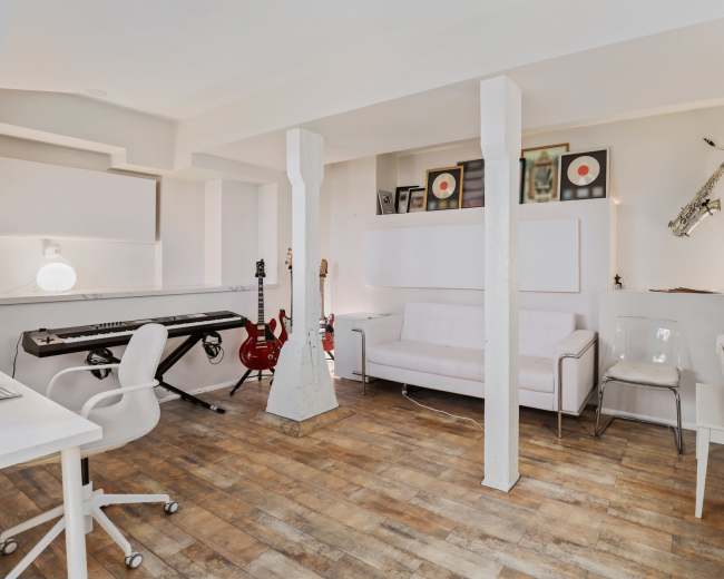 A modern home studio features a keyboard, electric guitars, a white couch, and a saxophone mounted on the wall, all arranged in a bright setting.