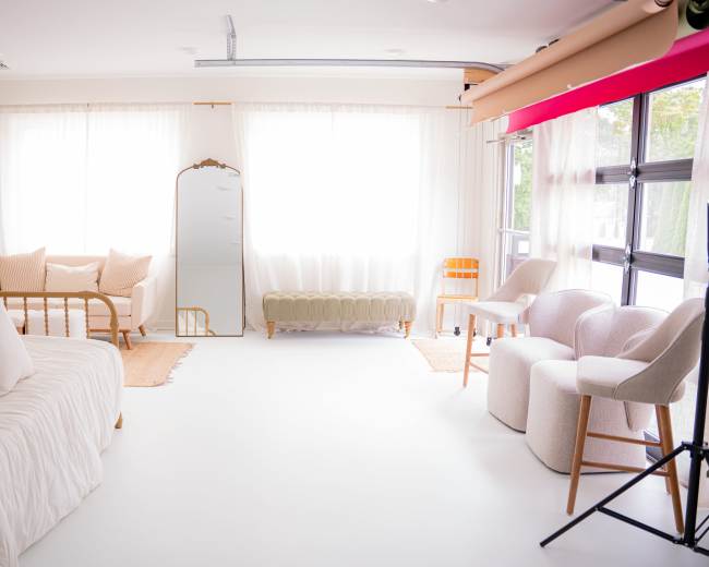 The image shows a bright, minimalistic interior space with a daybed, two armchairs, a mirror, and a green upholstered bench, all set against large windows with sheer curtains.