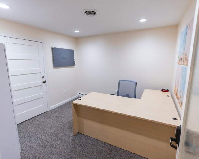 The image shows a small office with a light-colored desk, a chair, and a welcome sign on the wall, featuring neutral-colored walls and carpet.
