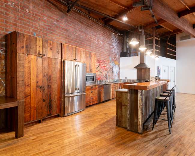 The image shows a spacious industrial-style kitchen featuring brick walls, wooden cabinetry, a stainless steel refrigerator, and a long bar with metal stools.