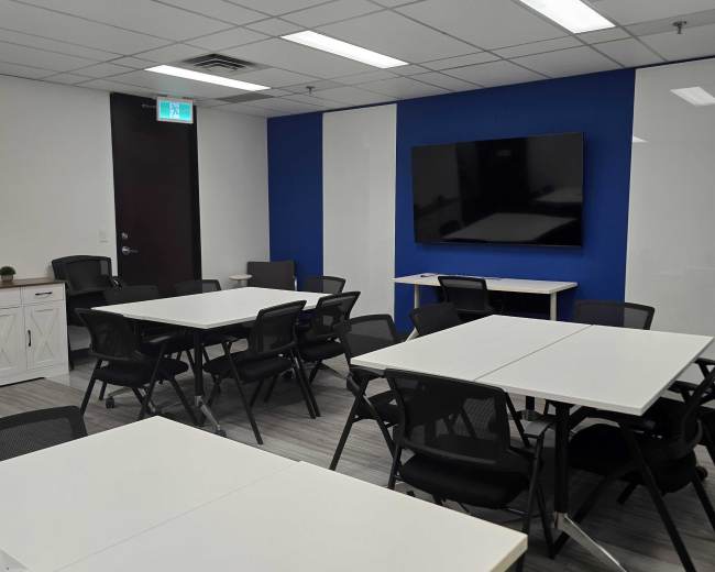 The image shows a modern conference room with multiple tables and black chairs arranged for meetings, featuring a large wall-mounted screen.