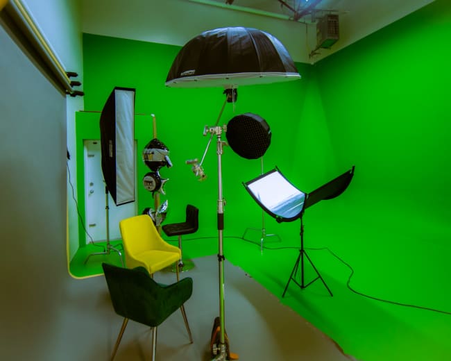 The image shows a photography studio set up with a green screen, lighting equipment, and two chairs in different styles.