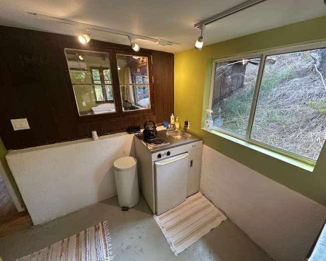 The image shows a small kitchen area with a sink, stove, and window overlooking a wooded landscape.