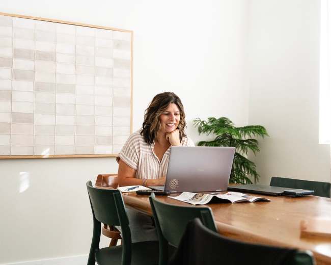 A woman with wavy hair sits at a wooden table with a laptop, smiling as she works in a well-lit room featuring a large art piece on the wall and a potted plant nearby.