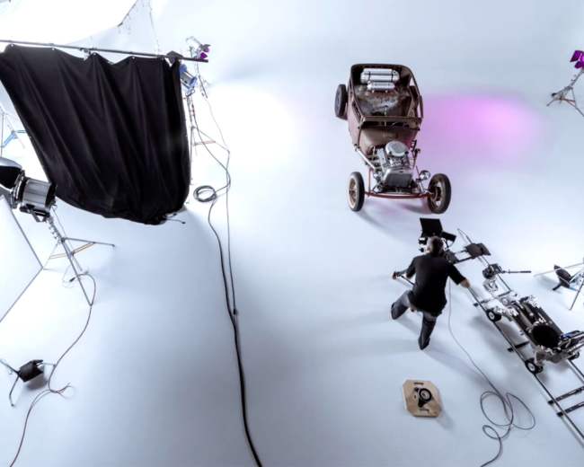 A studio set is arranged with various lighting setups, a black curtain, and a vintage car positioned on a light-colored floor.