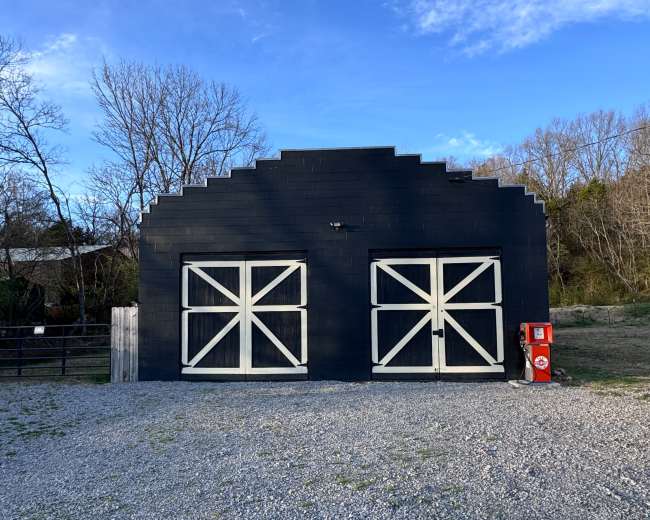 A large black barn with white doors stands against a clear blue sky, accompanied by a vintage gas pump on the side.