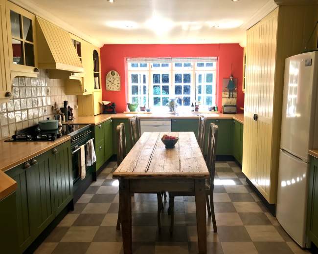 The image shows a spacious kitchen with green and yellow cabinets, a wooden dining table in the center, and a large window providing natural light.