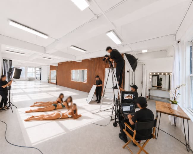 A photography studio features several people capturing images of two models posing on the floor, surrounded by various lighting equipment and a wooden backdrop.