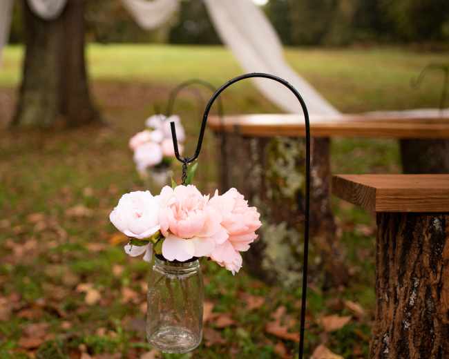 A glass mason jar filled with pink flowers hangs from a metal holder on a rustic wooden table in a grassy outdoor setting.