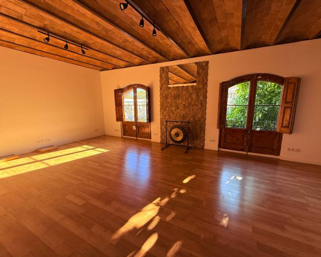 The room features wooden beams on the ceiling, a stone wall with a mirror, a large gong, and two open windows allowing natural light to enter.