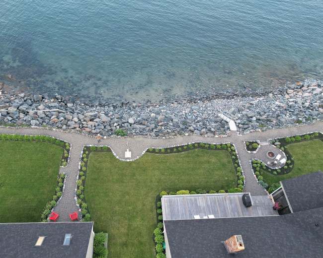 The image shows a coastal property with two neatly landscaped green lawns, bordered by a gravel path and rocky shoreline leading to the water's edge.