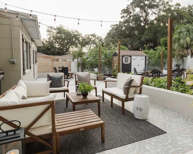 The outdoor area features a seating arrangement with white chairs and wooden tables on a patterned rug, surrounded by greenery and a small shed in the background.