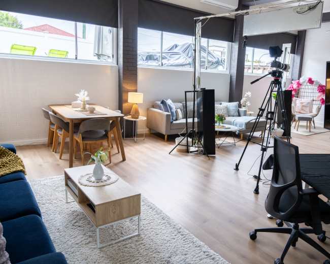 The image shows a modern studio space featuring a living area with a sofa and a dining table, equipped with cameras and a large screen for production purposes.