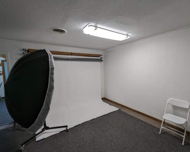 A photography studio with a white backdrop and a portable reflector on a stand, along with a single folding chair against a plain wall.