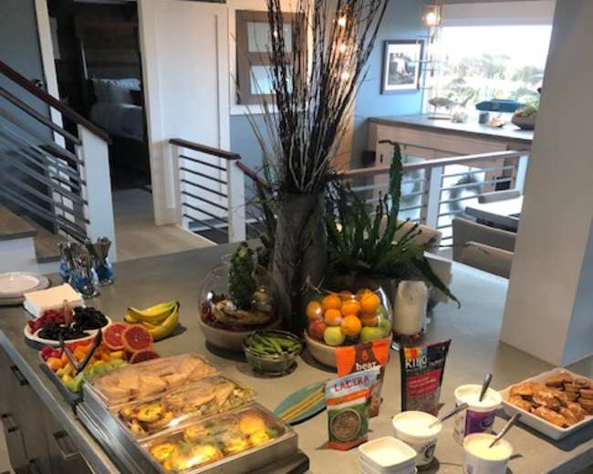 The image shows a buffet-style spread on a kitchen island, featuring a variety of fruits, pastries, and snacks, along with dips and beverages.