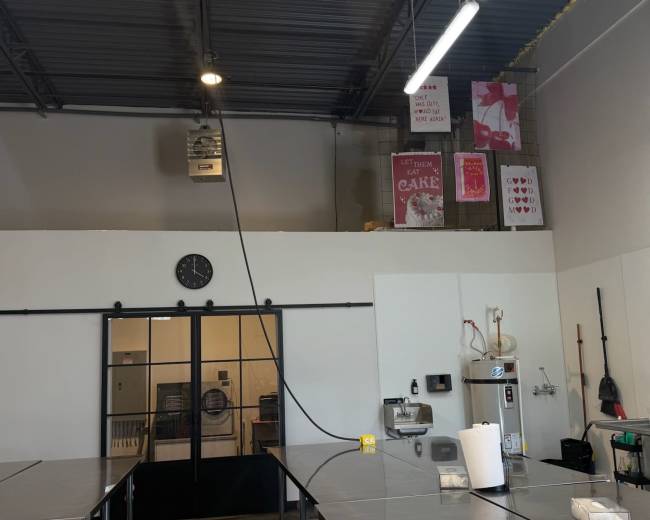 The image shows an interior view of a commercial kitchen with stainless steel countertops, a clock on the wall, and various kitchen equipment.