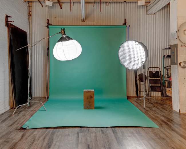 The image shows a photography studio setup with a green backdrop, a stool, and two lighting sources positioned on either side.