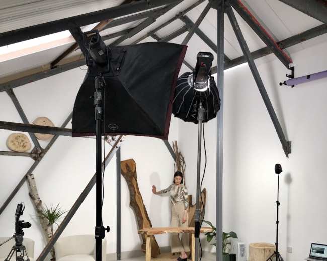 A person stands next to a wooden table in a studio setting equipped with photography lights and various props.