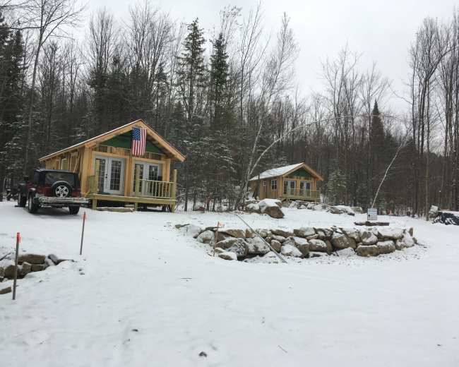 Two wooden cabins are situated on a snowy landscape surrounded by trees, with an American flag displayed on one of the cabins.