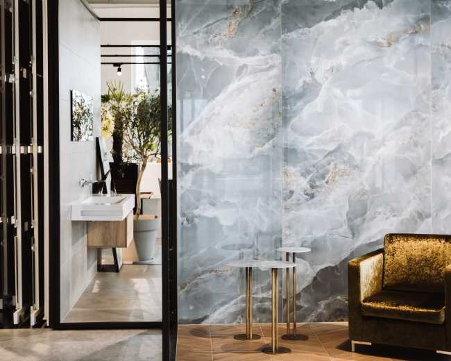 The image shows a modern interior with a gray marble wall, a small wooden floor area, a gold-accented chair, and a view of a sink and plants beyond a glass partition.