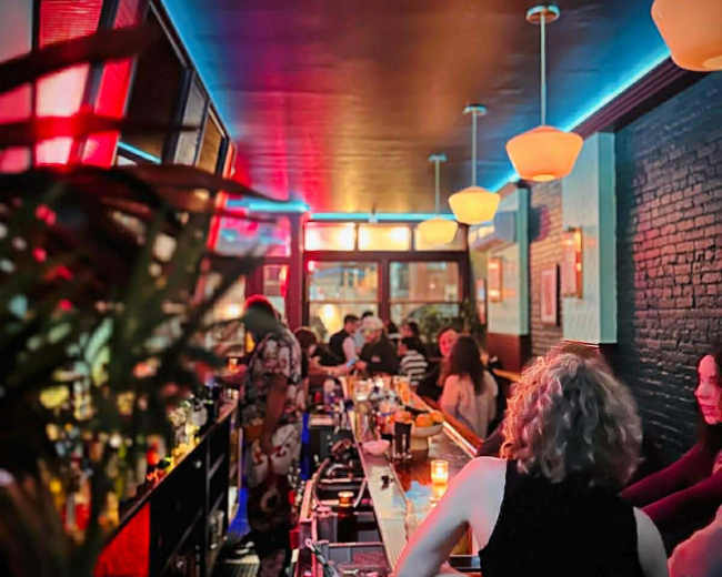 The image shows a dimly lit bar with patrons sitting at the counter and shelves lined with various bottles.