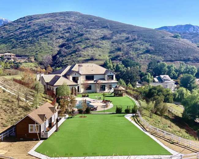 The image shows a large residential property with a green lawn, a swimming pool, and a playground, surrounded by hills and trees.
