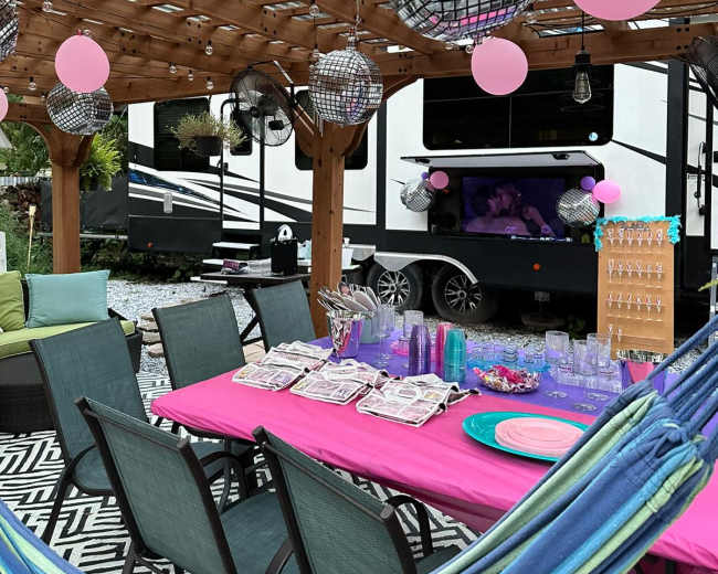 A backyard patio features a colorful table set for a party, adorned with balloons and disco balls, alongside an RV parked in the background.