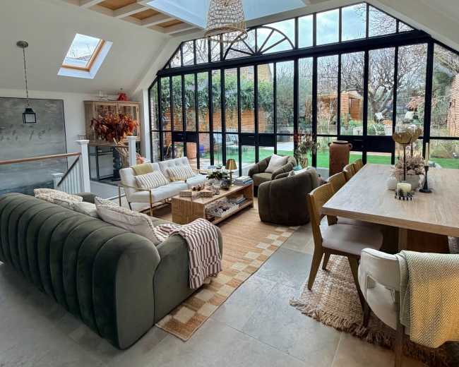 The image depicts a modern living room with large glass windows, a wooden ceiling, and a cozy seating arrangement, surrounded by indoor plants and a dining area.