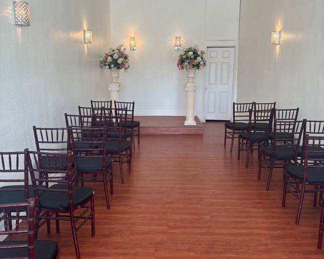 The image shows a narrow, well-lit room set up for a formal event, featuring rows of chairs facing a small stage with floral arrangements on pedestals.