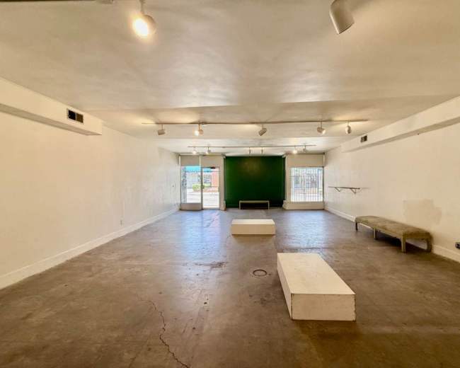 The image shows a spacious, empty room with concrete flooring, white walls, and a green wall at the far end, featuring two platforms and a bench.
