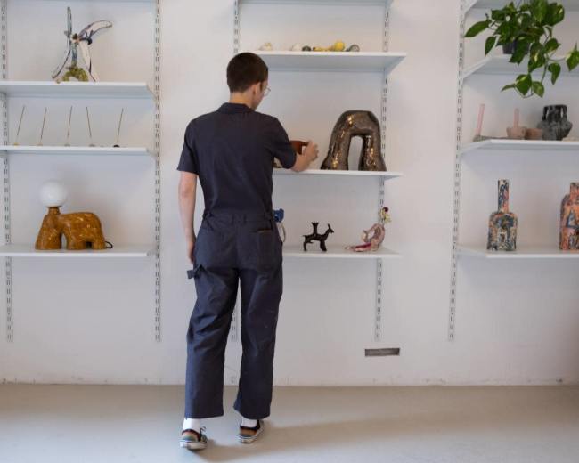 A person in a blue jumpsuit is arranging decorative objects on white shelves in a gallery or studio space.