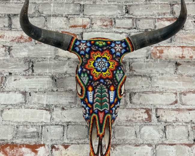 A colorful, intricately decorated bull skull is mounted on a textured brick wall.