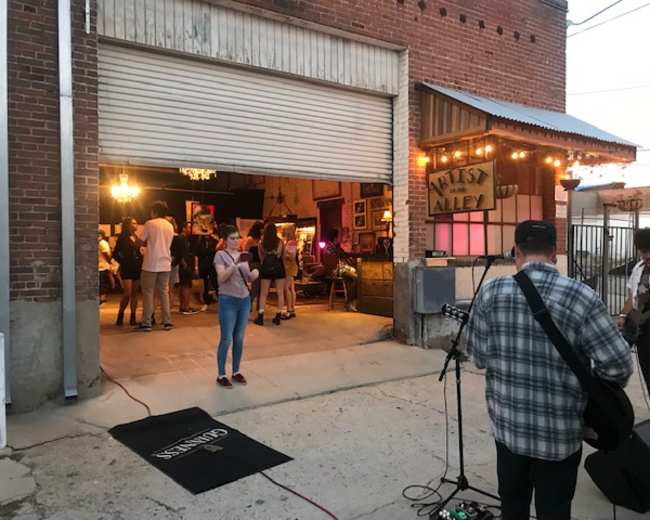 A group of people is gathered in an open alleyway for an outdoor music event, with a band playing inside a warehouse-like space adorned with colorful lights.