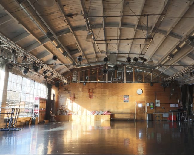 The image shows an empty gymnasium with a wooden floor, large windows, and overhead lighting.