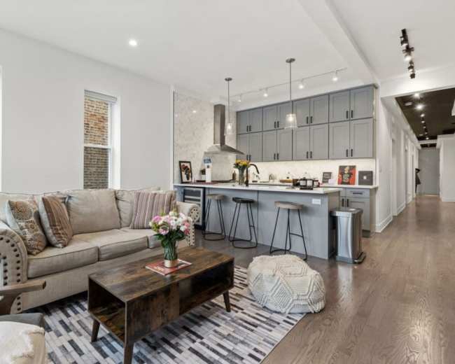 The image shows a modern living area featuring a beige sofa, a wooden coffee table, and a kitchen with gray cabinetry and a marbled backsplash.