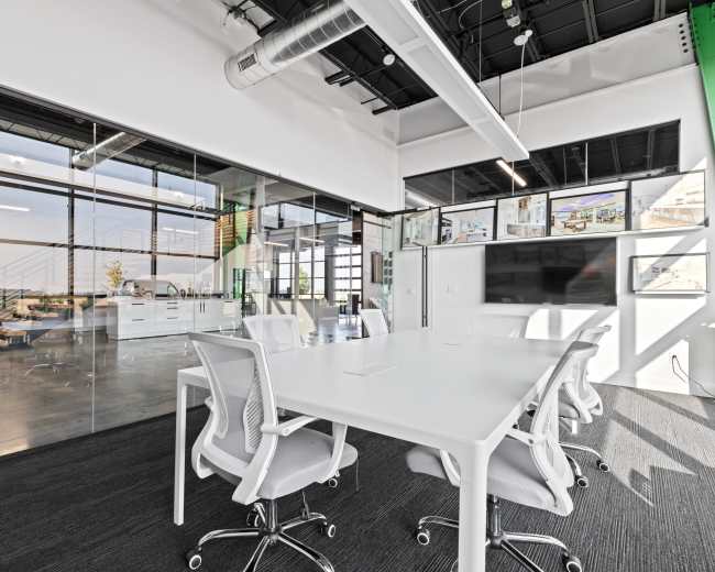 A bright conference room features a large white table surrounded by modern chairs, with glass walls that provide a view of a spacious office area.