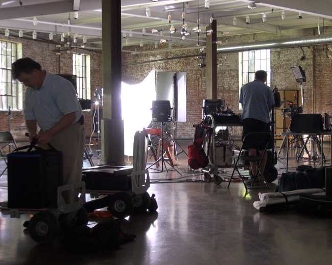 A film production crew sets up equipment in a spacious industrial workspace with large windows and exposed brick walls.