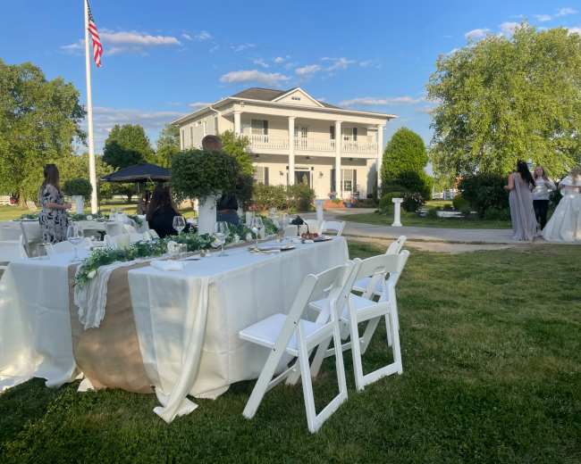 A wedding reception is set up outdoors with tables draped in white cloth, while guests mingle in front of a large, two-story venue surrounded by greenery.