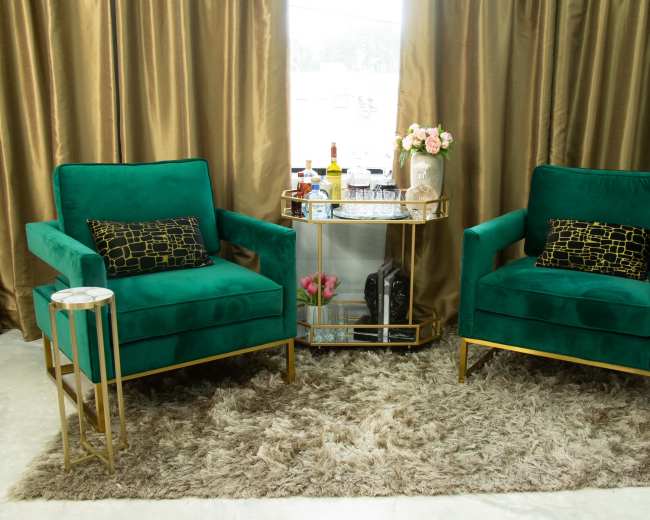 The image shows two green velvet armchairs with gold accents flanking a glass-top side table on a plush area rug in a room with golden curtains.