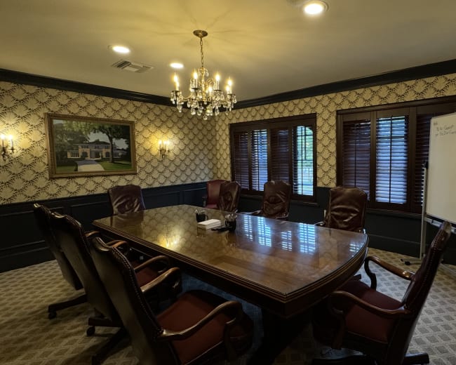 The image shows a well-furnished conference room featuring a large wooden table, leather chairs, a whiteboard, and windows with wooden blinds.