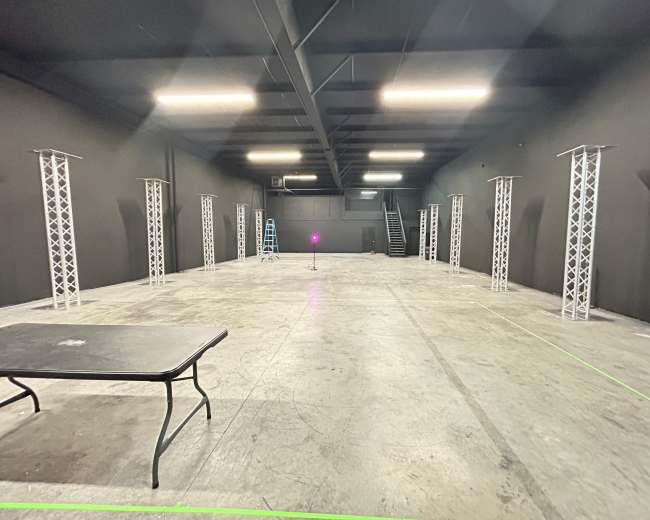 The image shows an empty industrial space with a concrete floor, black walls, and several tall, white truss structures arranged around the room.