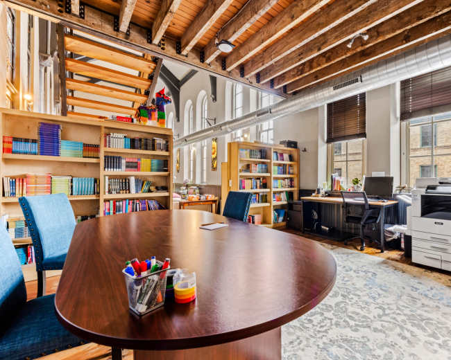 The image shows an office space featuring a large wooden table, bookshelves filled with books, a desk with a computer, and a staircase leading to a loft area.