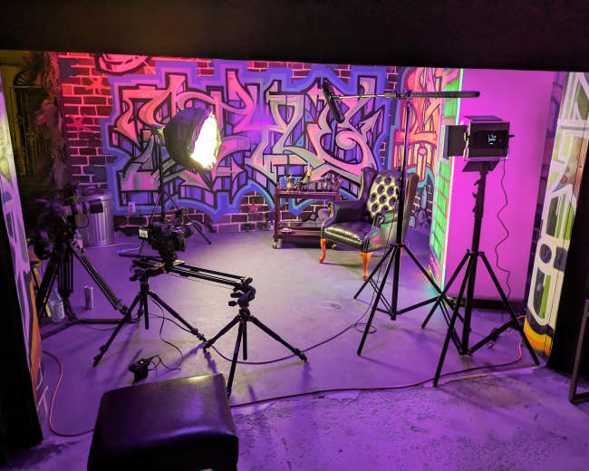 The image shows a photography studio setup with various lights and cameras positioned around a black chair in front of a colorful graffiti backdrop.