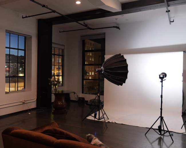 The image shows an interior space with a large window, a brown couch, a photography lighting setup, and a reflective mirror leaning against the wall.