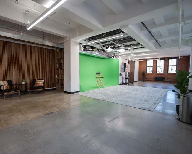 The image shows a spacious studio with a wooden accent wall, a green screen area, and a seating area that includes two chairs and a decorative plant.